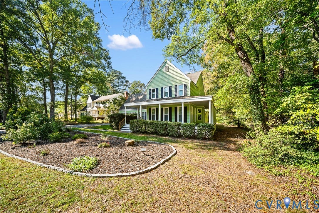 1608 Queens Point Drive, Henrico, VA 23238 Two-story exterior featuring light green horizontal siding, dark shutters, a covered wrap-around porch, and stone-bordered garden beds situated among mature trees