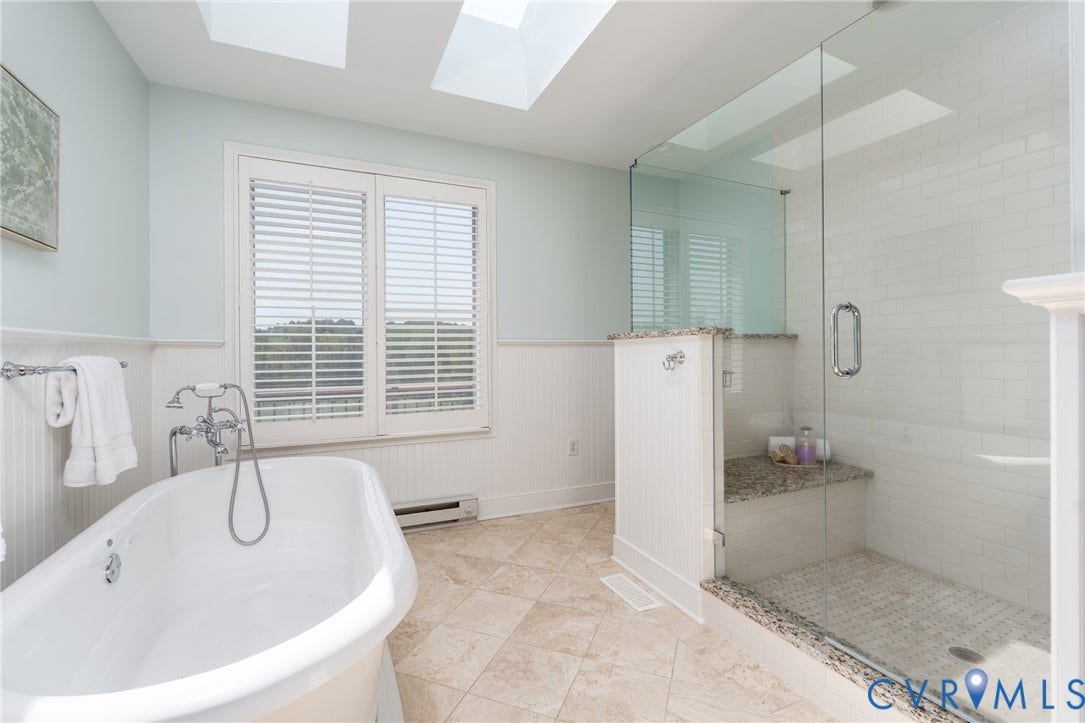 Freestanding soaking tub with chrome fixtures, glass-enclosed shower with subway tile, built-in shower bench with stone surface, skylights, and plantation shutters