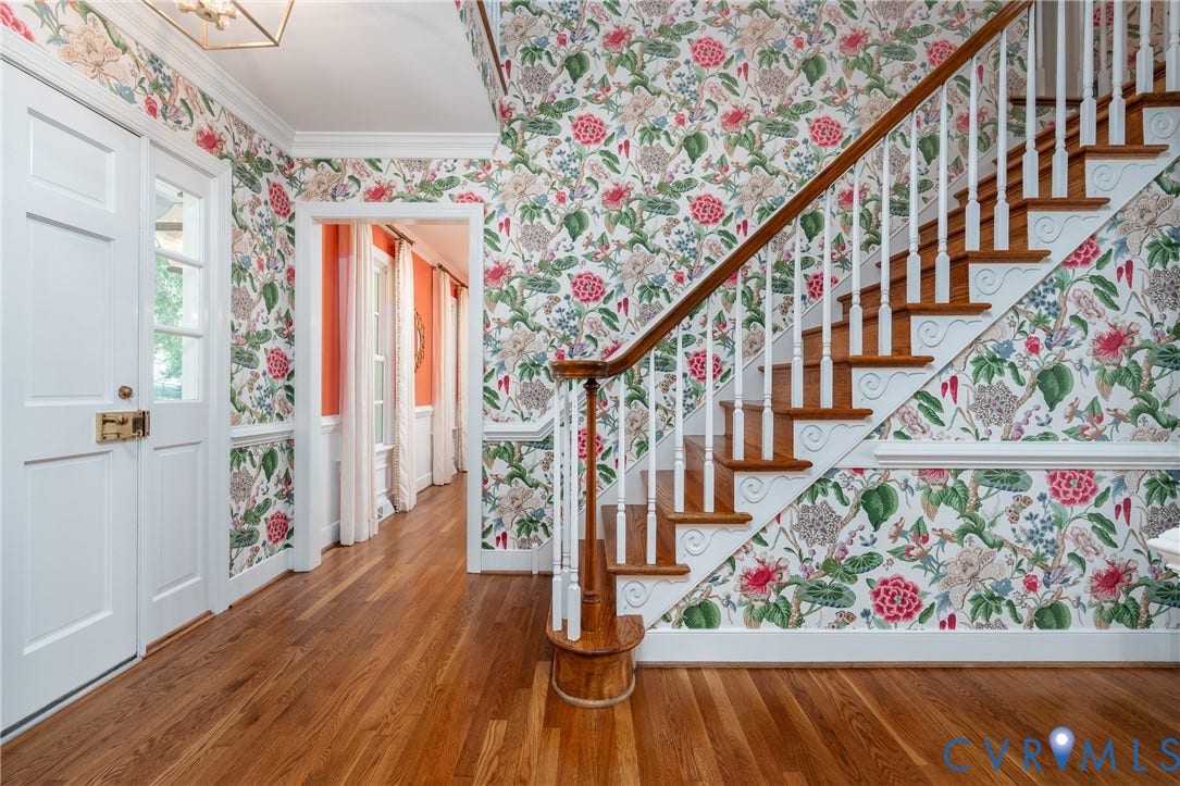 Hardwood flooring, decorative floral wallpaper, white paneled door with brass hardware, open archway, and a staircase with white balusters and wood treads