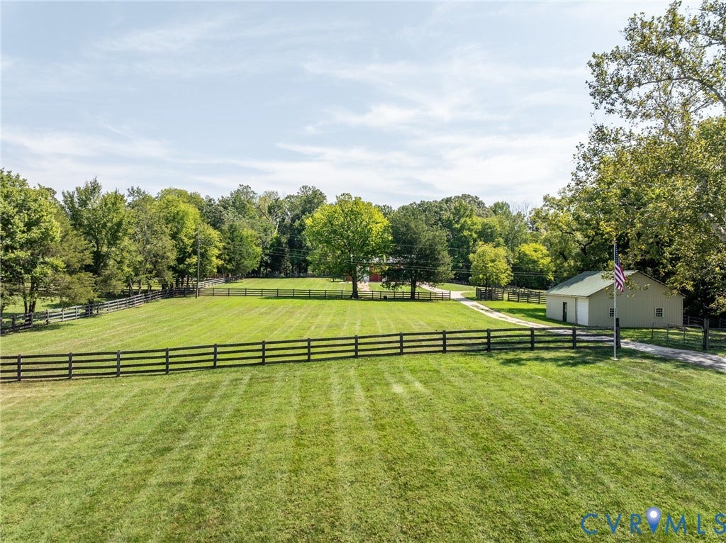 Expansive green lawn with dark wood fencing, mature trees, a gravel driveway, and a detached structure with a green roof