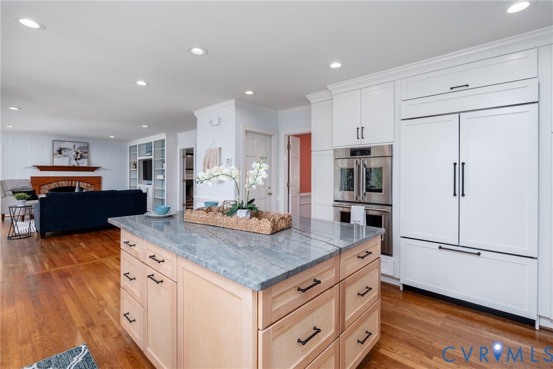 Kitchen island with light wood cabinetry and stone countertop, integrated paneled refrigerator, built-in stainless steel double ovens, white upper and lower cabinetry, and wood-finish flooring