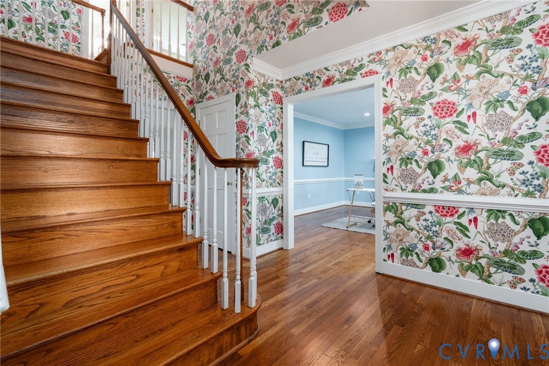 Hardwood flooring throughout, a staircase with white spindles and a wood handrail, crown molding, and botanical-patterned wallpaper