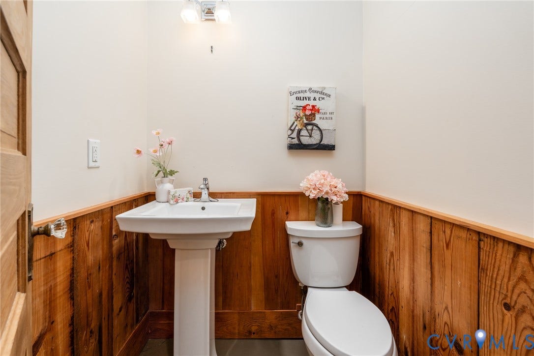 Wood-paneled wainscoting, pedestal sink, chrome faucet, white toilet, and overhead vanity lighting