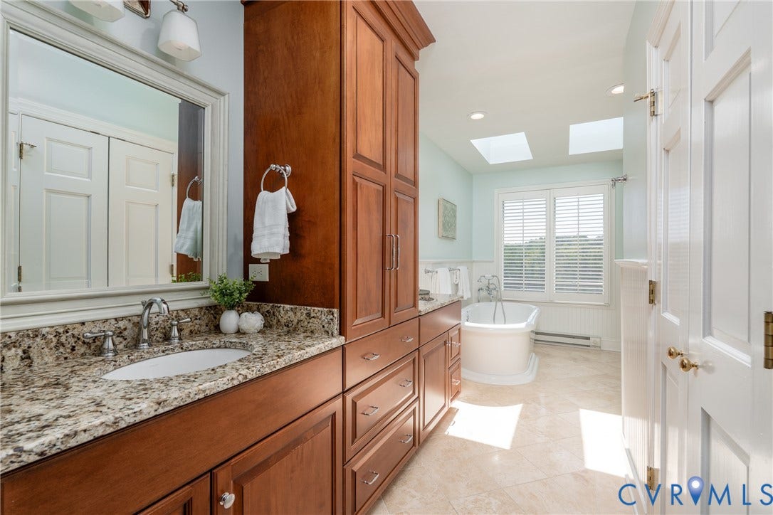Expansive bath featuring a freestanding tub, dual skylights, wood-finish cabinetry, a single vanity with stone countertop, and tile flooring