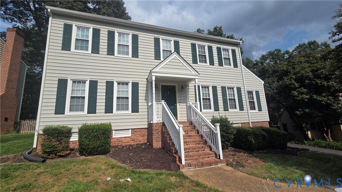 Two-story colonial exterior with light-colored siding, brick foundation, a covered entry porch, and dark shutters