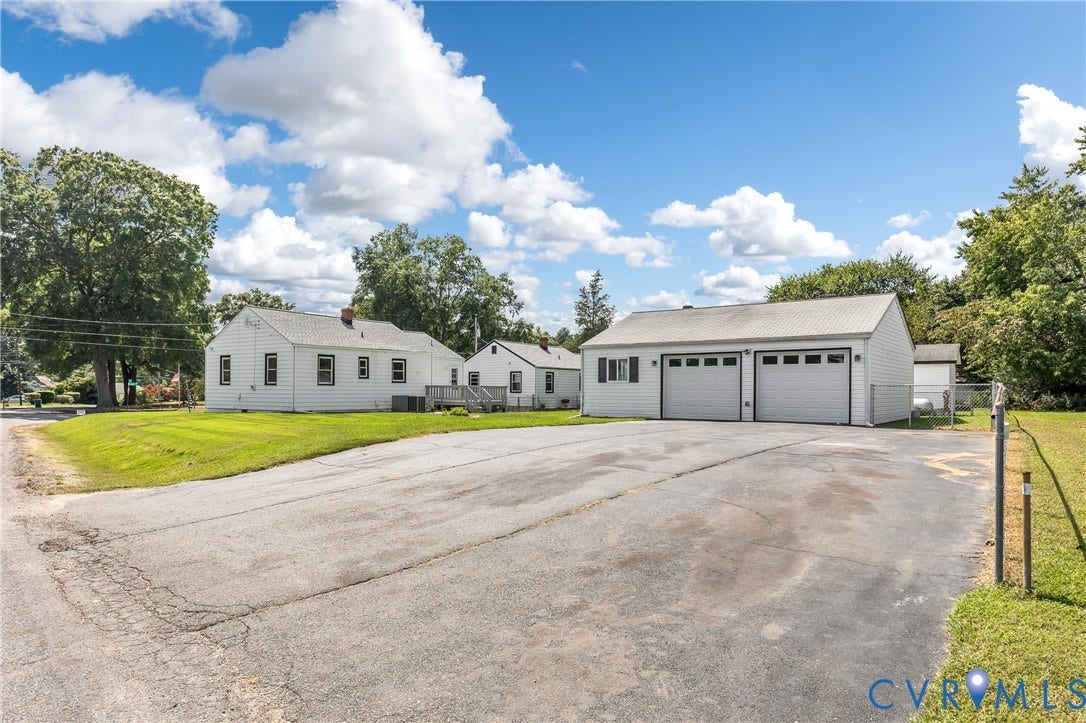 White exterior with dark trim windows, attached two-car garage, expansive paved driveway, and a secondary structure