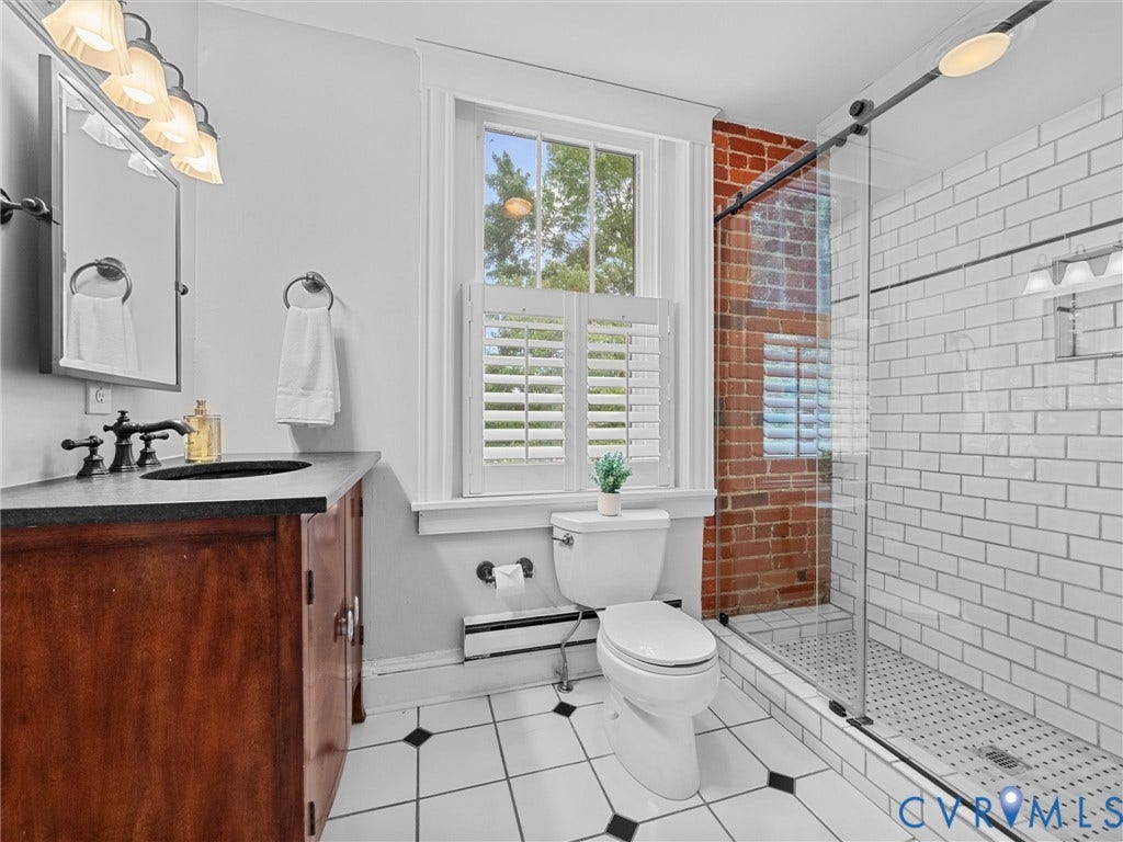 3403 W Franklin Street, Museum District, Richmond, VA 23221 Bathroom featuring a glass-enclosed shower with white subway tile, exposed brick wall, wood vanity with dark countertop, and a window with plantation shutters