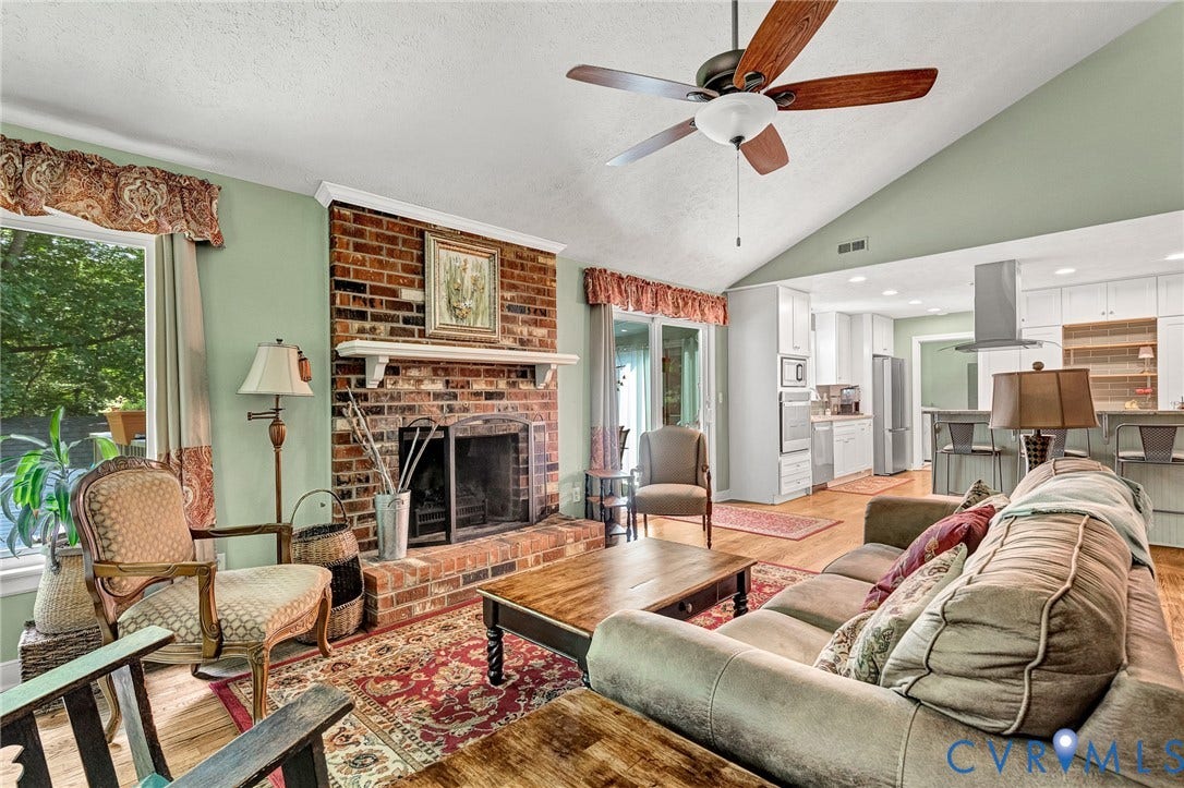 Vaulted ceiling with a ceiling fan, brick fireplace with a mantel, wood-finish flooring, and a kitchen with stainless steel appliances