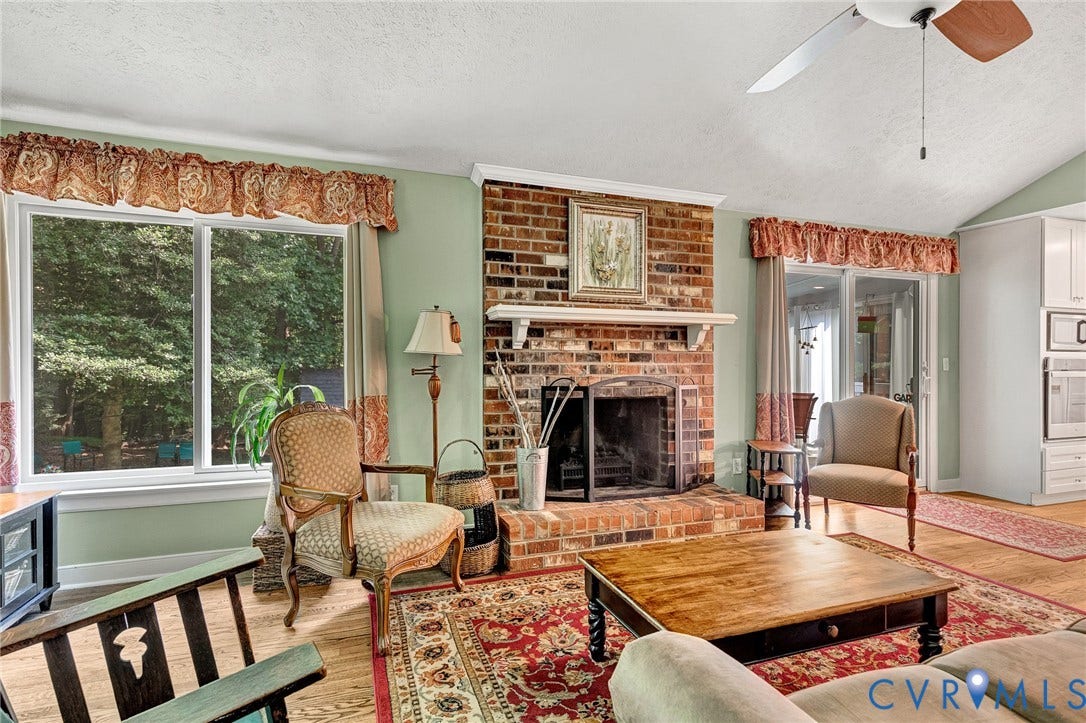 Brick-clad fireplace with mantel, vaulted ceiling with ceiling fan, and large windows