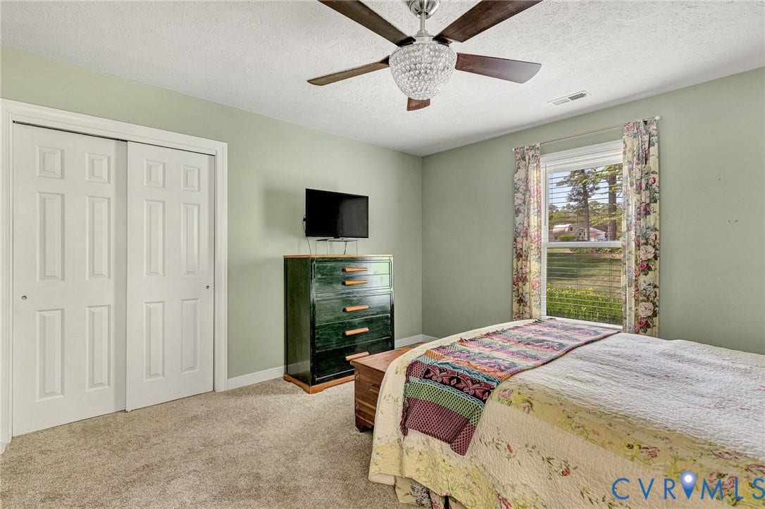 Plush carpet flooring, ceiling fan with chandelier lighting, and a single window with blinds