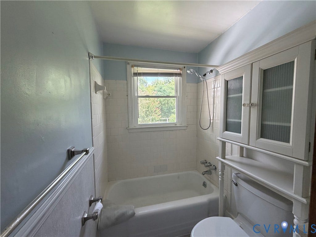 Built-in storage cabinet with fluted glass doors, full bathtub with shower, white subway tile surround, and a window with natural light