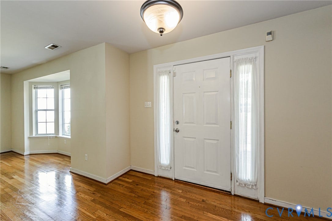 Hardwood flooring throughout, a front entry door with sidelights, a ceiling-mounted light fixture, and a bay window in an adjoining area
