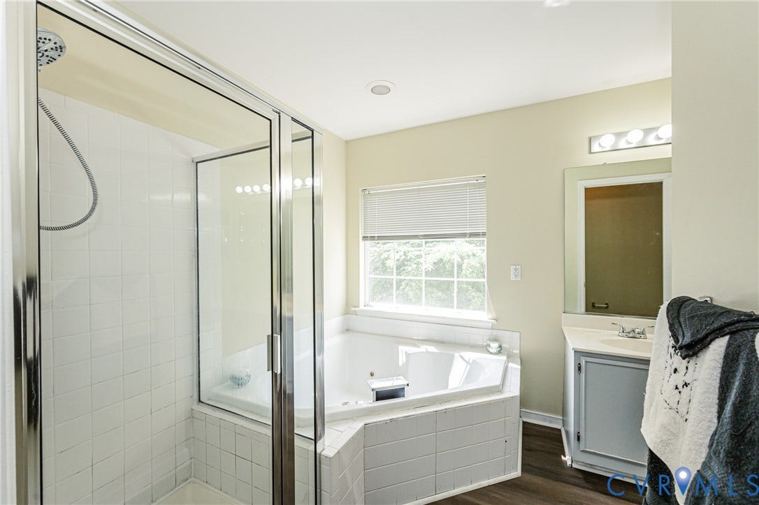 Spa-inspired primary bathroom featuring a frameless glass-enclosed shower with subway tile surround, a soaking tub positioned beneath a transom window, dual vanity with white cabinetry and integrated sink, and wood-finish flooring throughout
