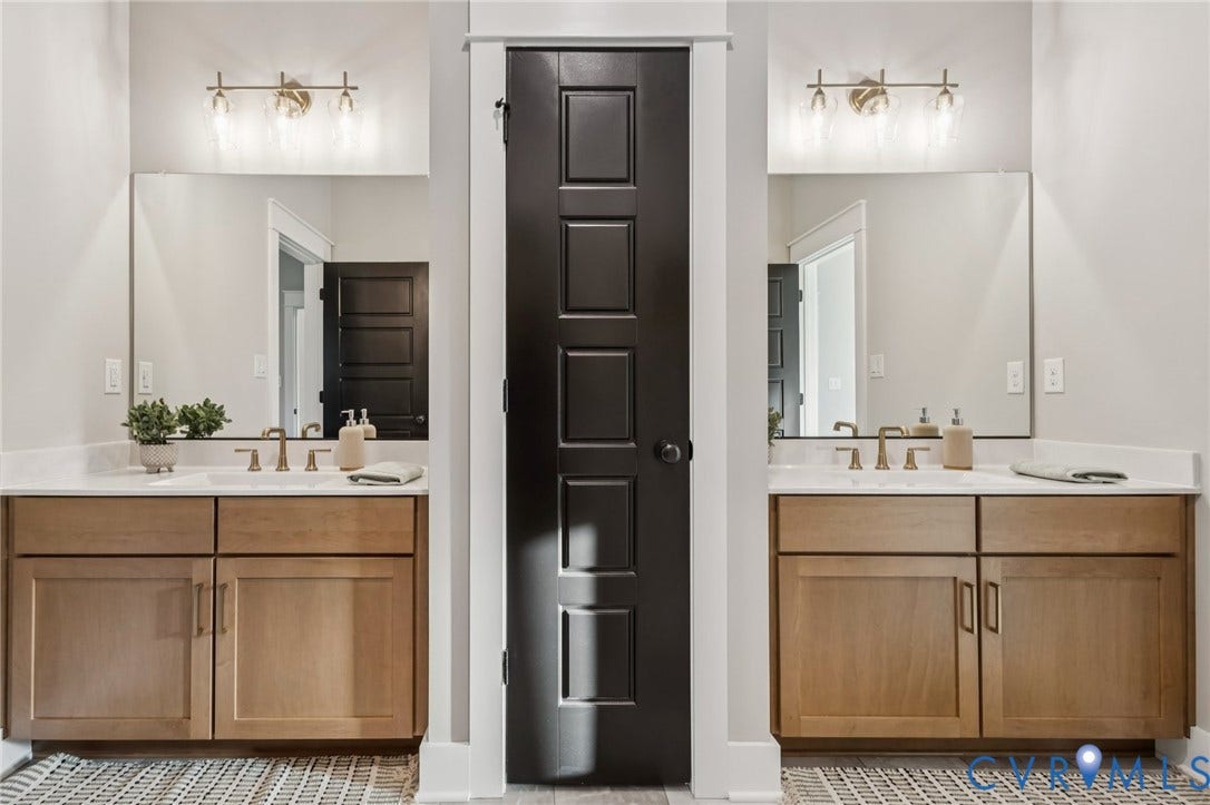 Lot 5 Olivet Church Road, Olivet Church Estates, New Kent, VA 23124 Dual vanity bathroom featuring wood-finish cabinetry with paired sinks, brushed gold hardware and sconces, large framed mirrors, and patterned tile flooring