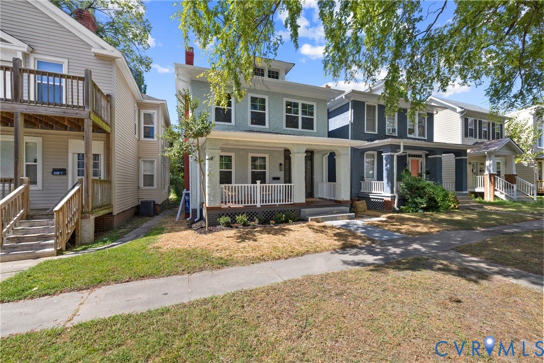 Two-story exterior with light blue stucco siding, a covered front porch with white railings, and a front yard with a concrete sidewalk