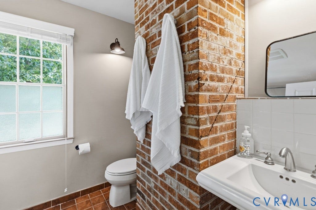 2416 Mountainbrook Drive, Crown Grant, Henrico, VA 23233 Exposed brick accent wall, white pedestal sink with dual-handle faucet, white tiled backsplash, rectangular framed mirror, and a window with white louvered blinds