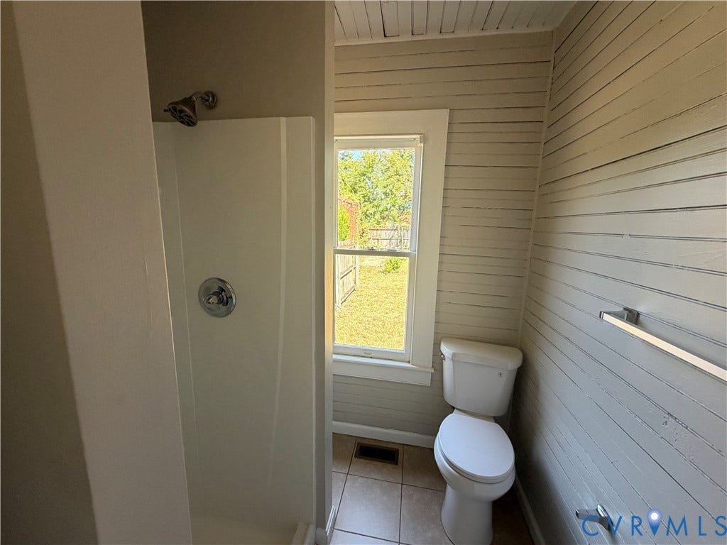 715 High Street, Petersburg, VA 23803 Tiled flooring, white shower enclosure with chrome fixtures, window with yard exposure, white toilet, and horizontal wood-finish wall paneling