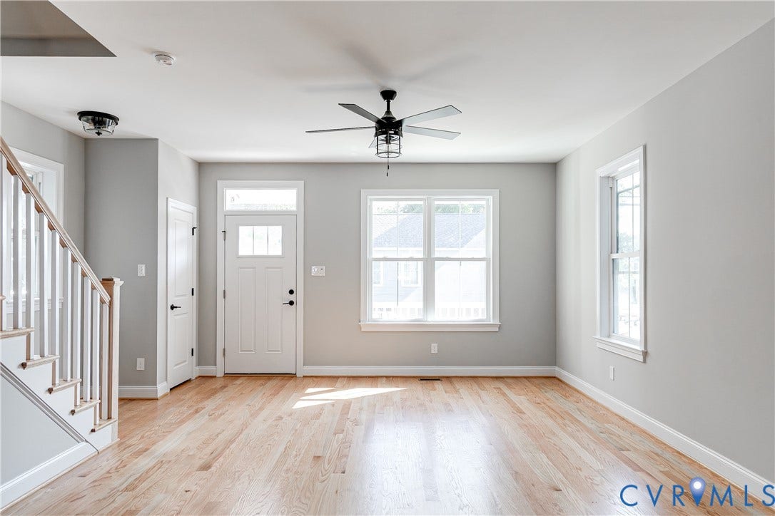 Hardwood flooring, white paneled entry door with transom and side window, white framed windows, ceiling fan with integrated lighting, and a staircase with white balusters and wood handrail