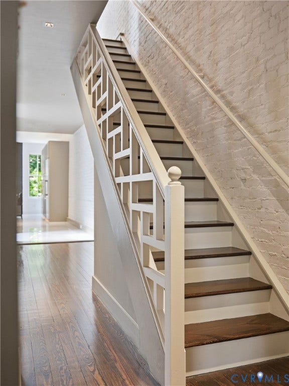 Hardwood flooring, painted brick accent wall, staircase with geometric balusters, dark wood stair treads, and recessed lighting