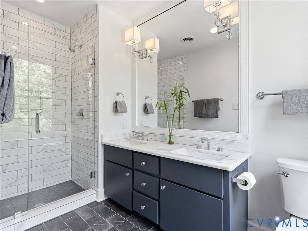 Glass-enclosed shower with subway tile surround, dark tile flooring, and a stone-topped double vanity with dual sinks