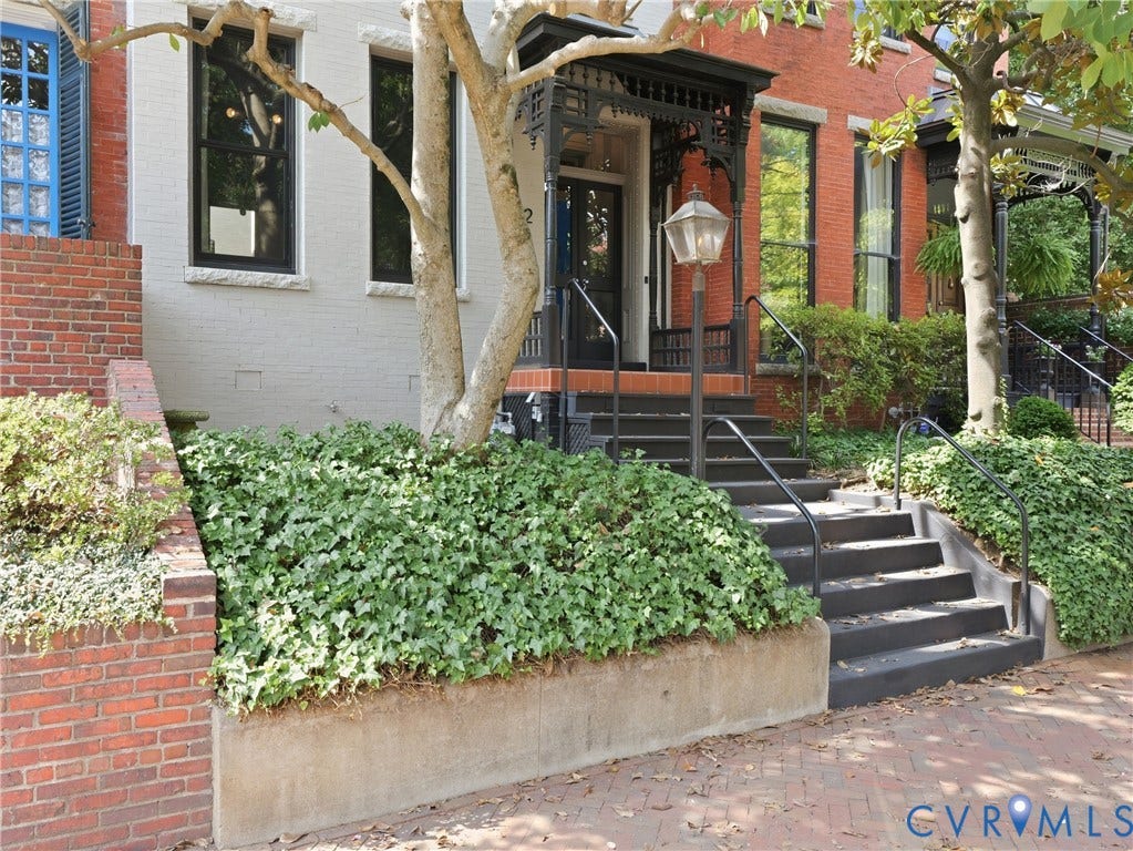 Brick exterior with black wrought iron railing, black exterior entry light fixture, and mature landscaping