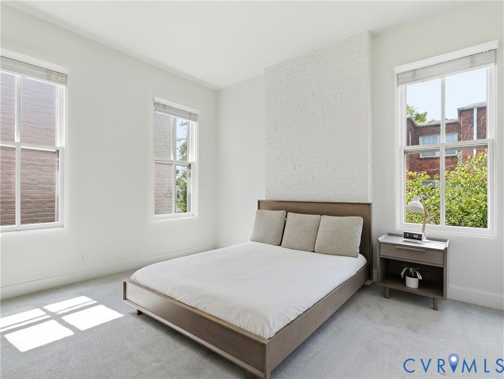 White painted brick accent wall, two large windows, light grey carpeting, and light-toned walls