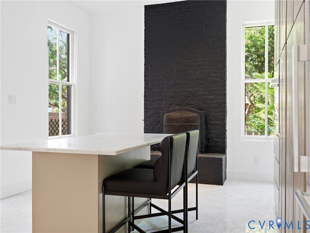 White countertop island, polished flooring, white walls, black brick fireplace, and tall windows