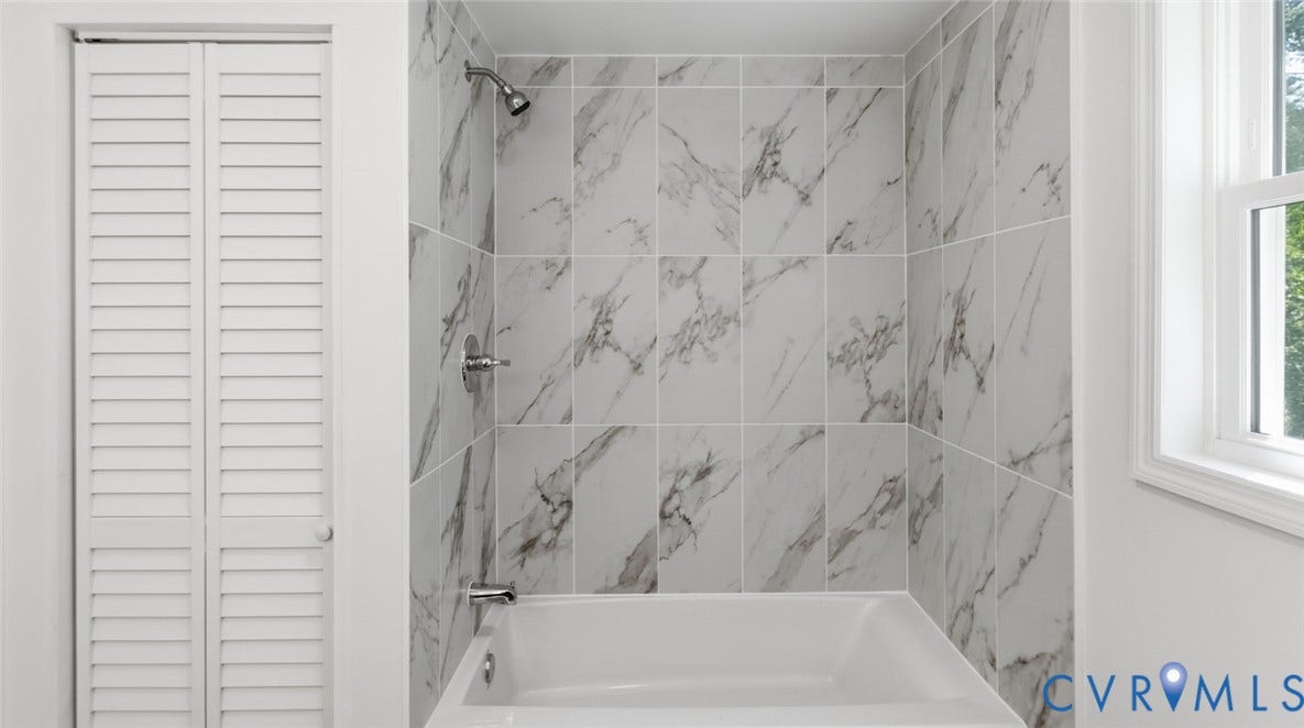 White bathtub with chrome fixtures, large format white tile shower surround, and a louvered bifold door