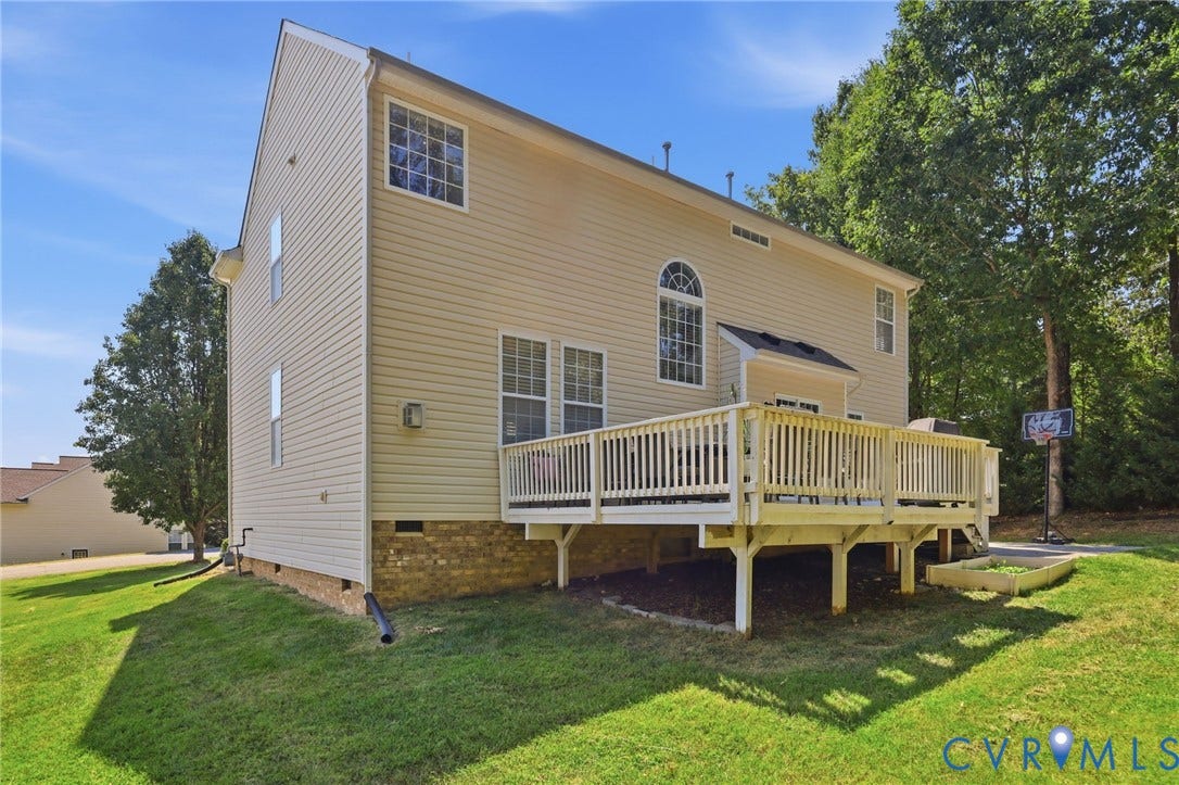 Expansive rear deck, brick foundation, light-colored siding, arched window, and mature trees surrounding the property