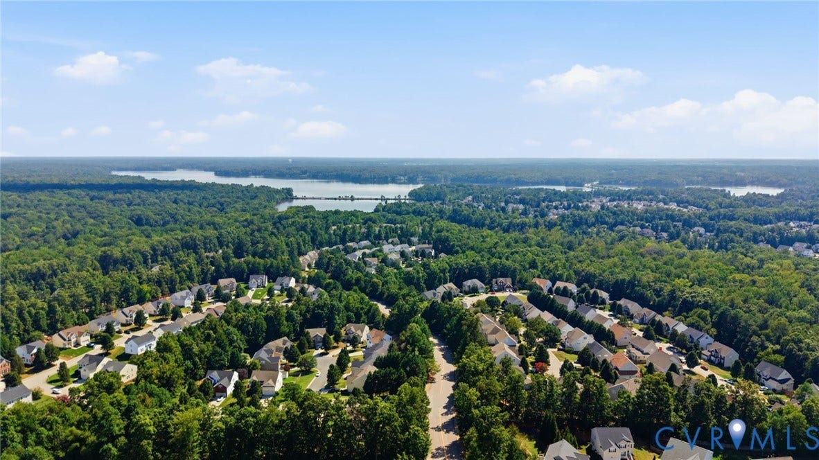 Expansive residential development surrounded by mature trees, featuring a serene lake and distant water bodies