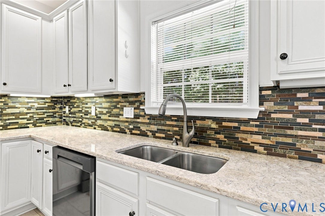 White cabinetry with dark hardware, mosaic tile backsplash, double basin stainless steel sink, stainless steel gooseneck faucet, and light-toned countertop