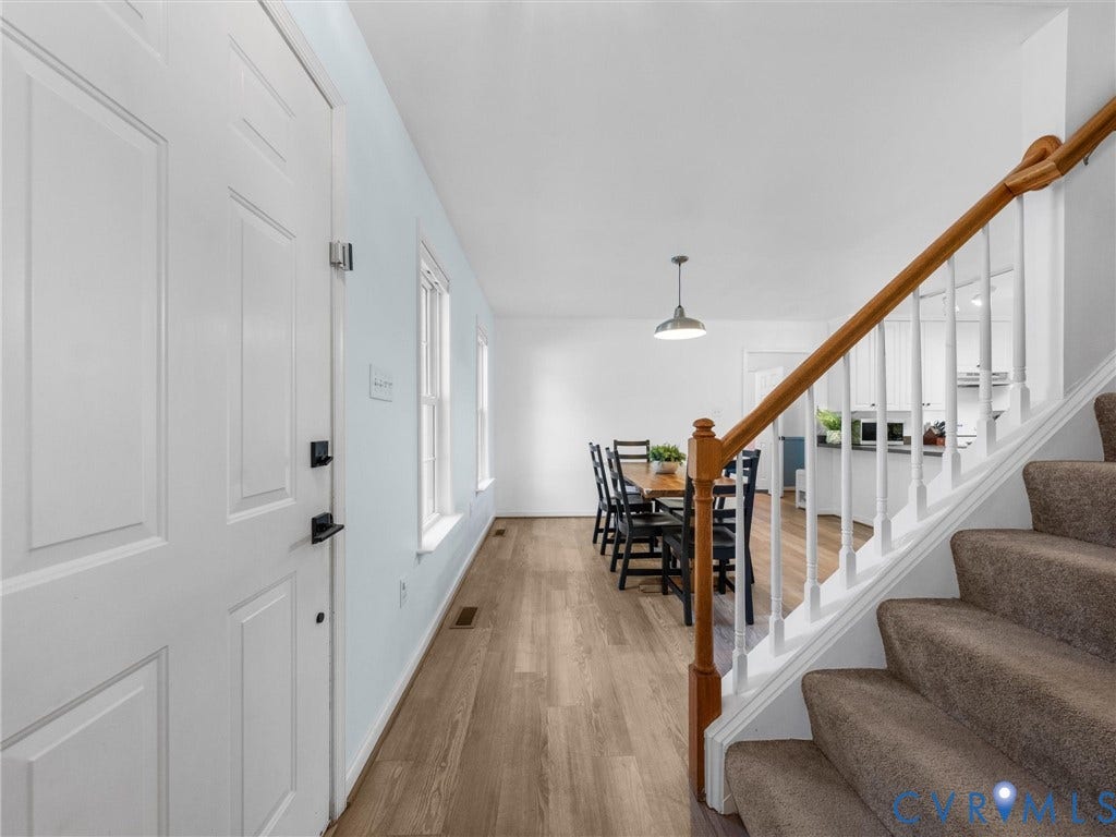 Panel door with black hardware, wood-finish flooring, two windows, and a carpeted staircase with wood handrail