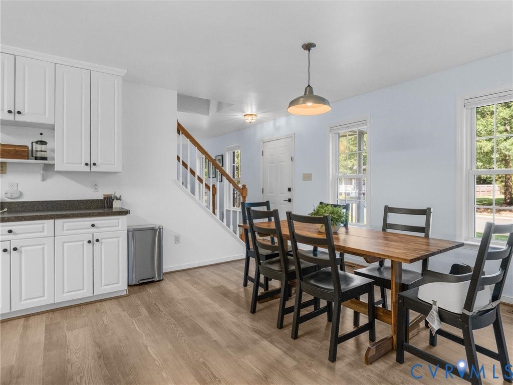 Light wood-finish flooring throughout, white shaker-style cabinetry, dark countertops, white double-hung windows, and a built-in staircase with wood railing