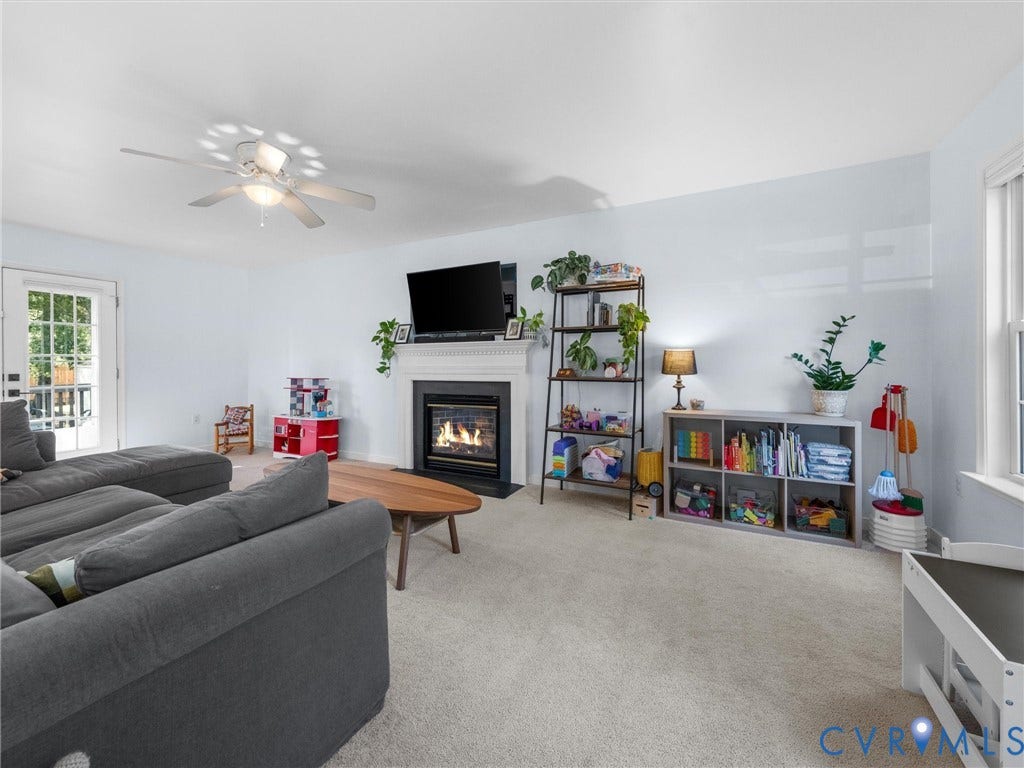 Carpeted living space featuring a fireplace with a white mantel, ceiling fan with integrated lighting, and a glass-paned door