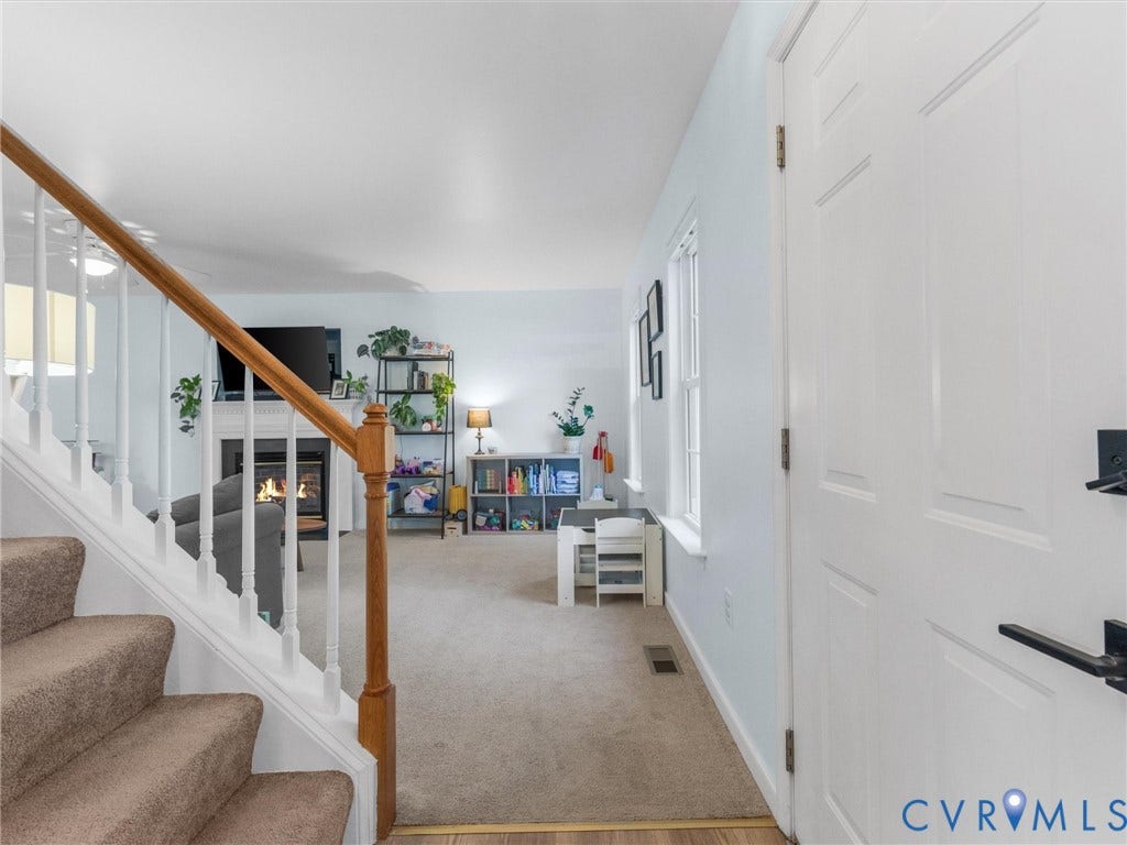 Carpeted staircase with wood railing, leading to a living area featuring a fireplace with white mantle and hearth