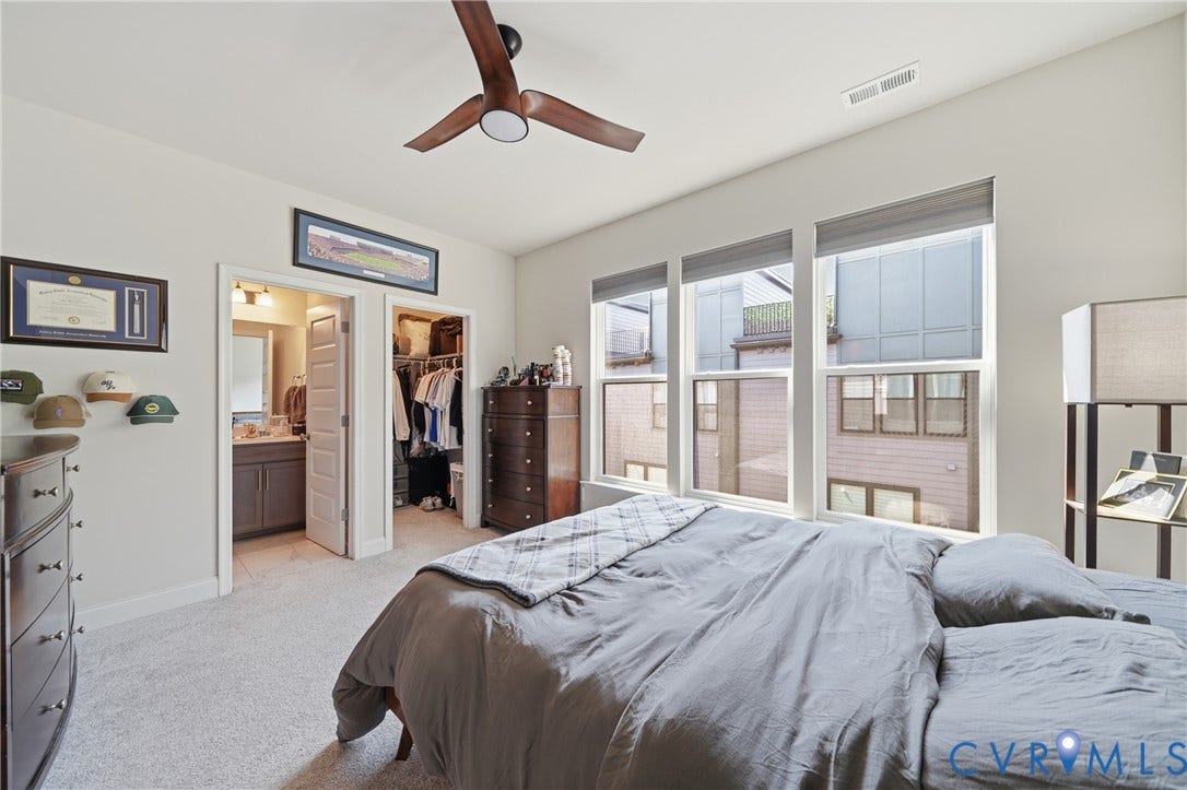 Plush carpeted flooring, three large windows with roller shades, and a wood-finish ceiling fan