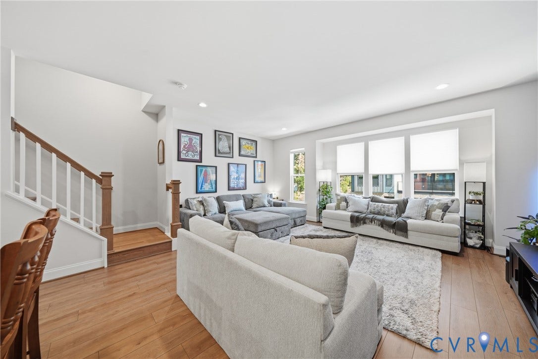Bright, open-concept living area showcasing hardwood flooring throughout, expansive windows with natural light, neutral upholstered seating arrangements, built-in shelving and media cabinetry, and a floating staircase with wood railings and white balusters