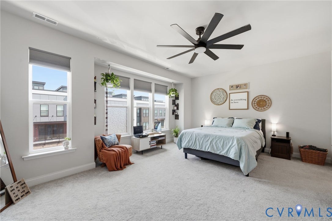 Spacious room with light gray carpeting, tall windows with roller shades, and a multi-blade ceiling fan