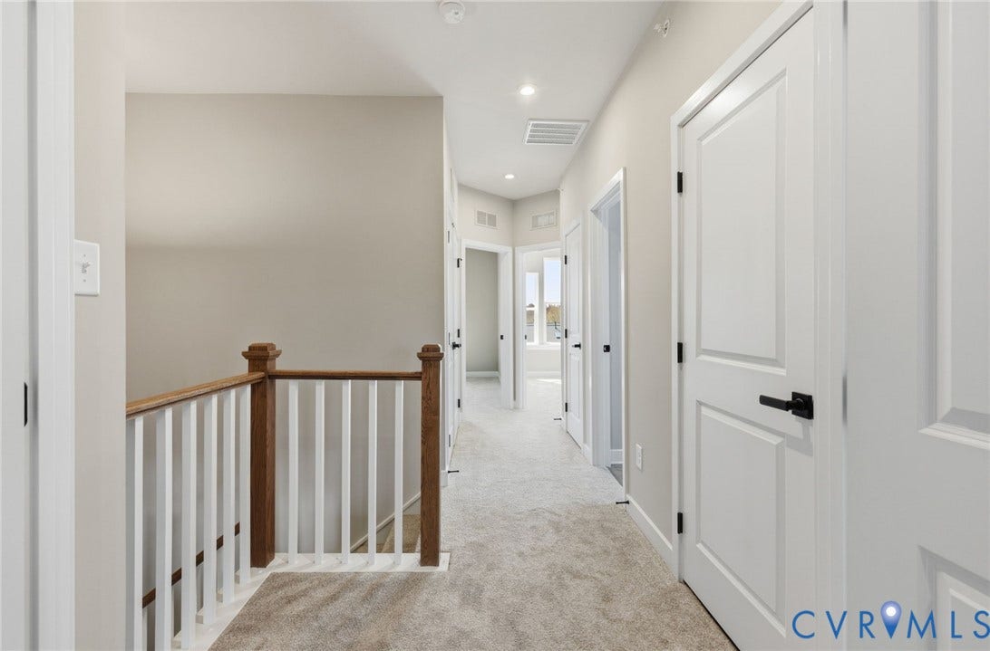 Hallway featuring light neutral wall paint, recessed lighting, carpet flooring, wood banister with white spindles, and white paneled doors with matte black hardware