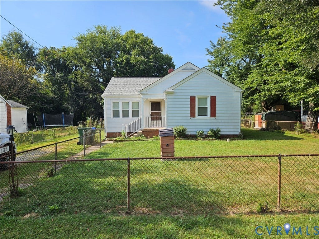 White siding exterior, front porch with railings, red window shutters, chain-link front fencing, and mature trees