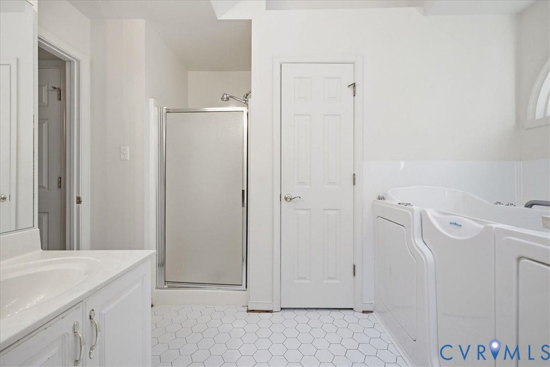 White vanity cabinet with integrated sink, chrome hardware, glass-enclosed shower, white hexagon tile flooring, and a walk-in bathtub