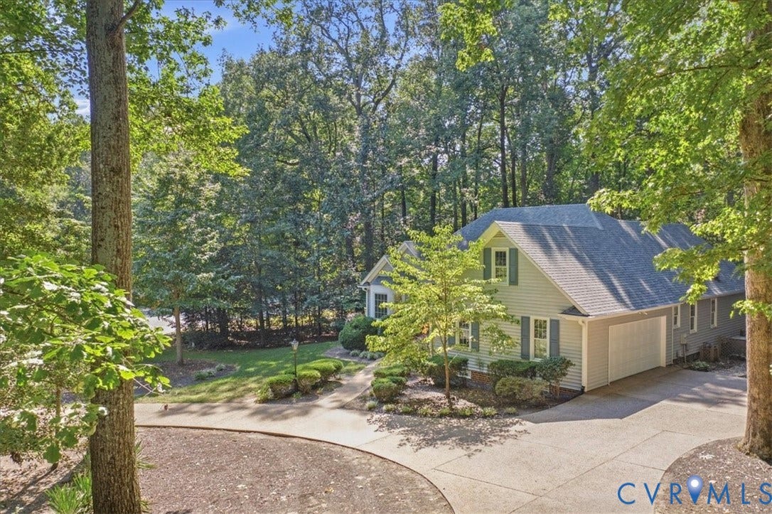 Siding exterior with dormer windows, attached garage, and a circular concrete driveway, surrounded by mature trees and landscaping