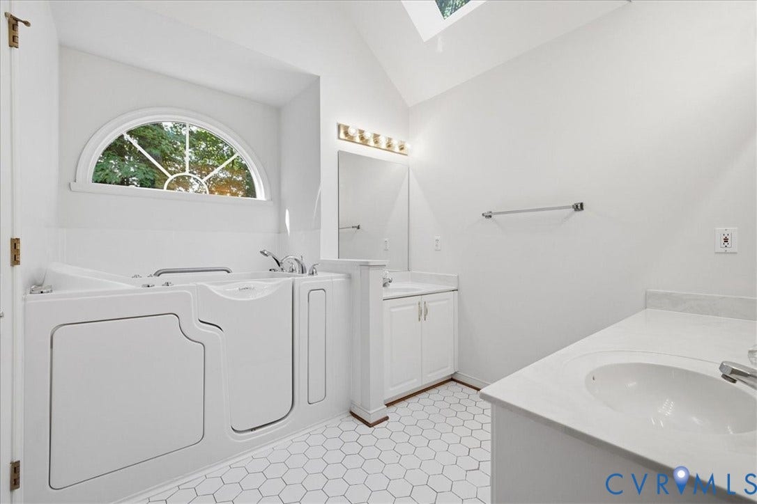 Walk-in tub with chrome fixtures, arched window, skylight, dual vanity with white cabinetry, and hexagonal tile flooring