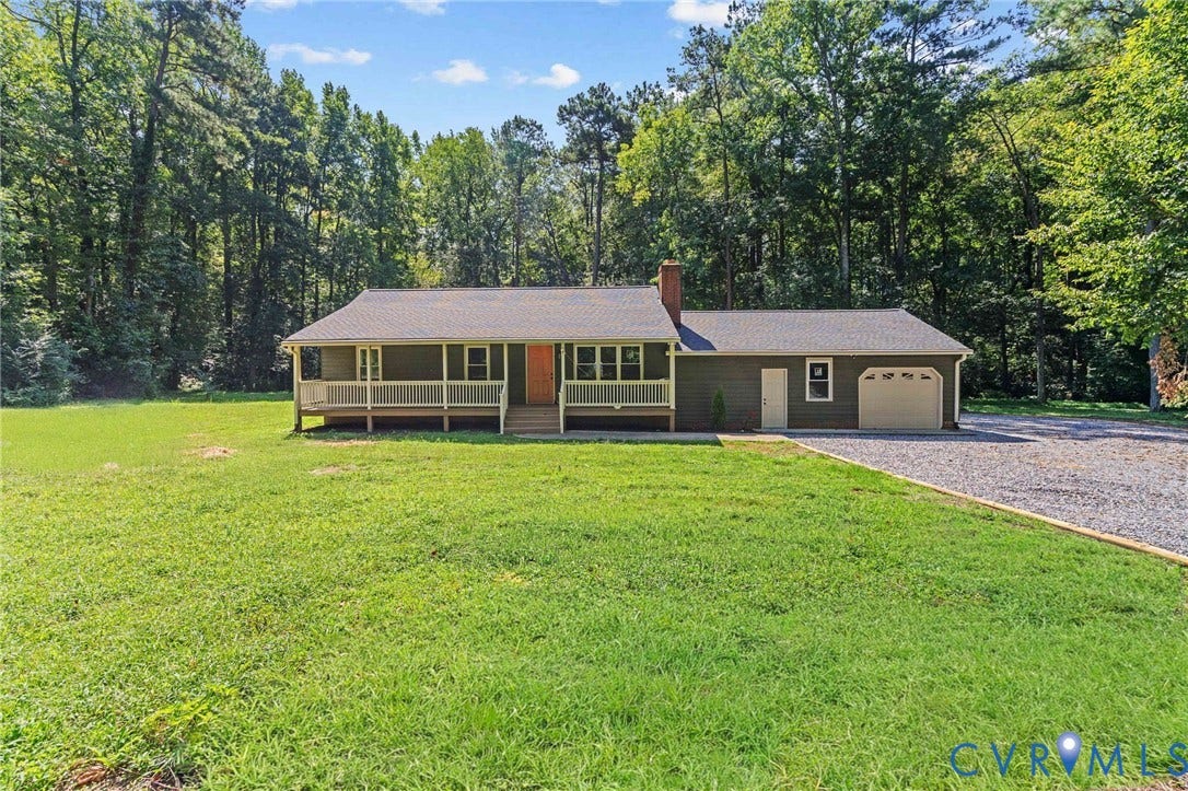 Ranch-style exterior with a covered front porch, attached garage, and gravel driveway, set against a wooded backdrop