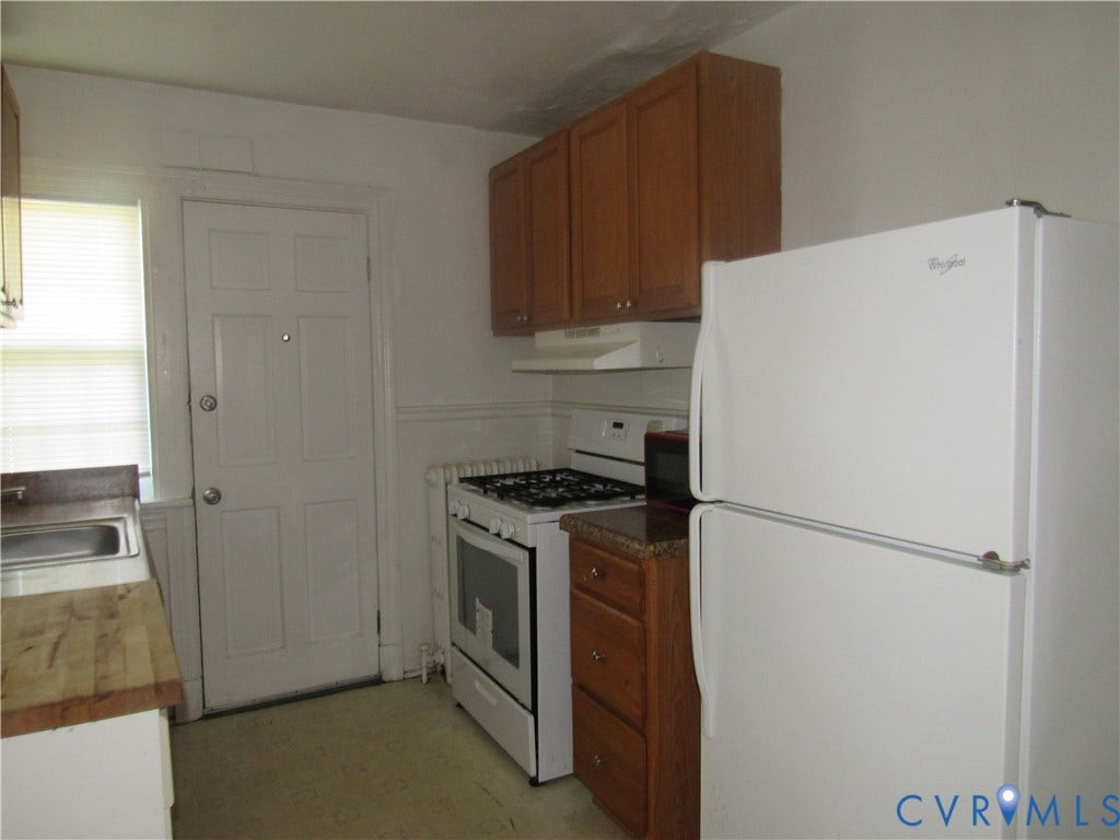 Full-size white refrigerator, gas range with range hood, wood-finish cabinetry, and a window with blinds