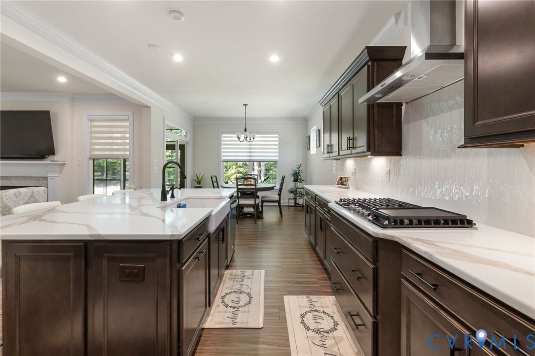 Expansive kitchen island with a farmhouse sink, integrated gas cooktop with a range hood, dark wood-finish cabinetry, and light-colored countertops