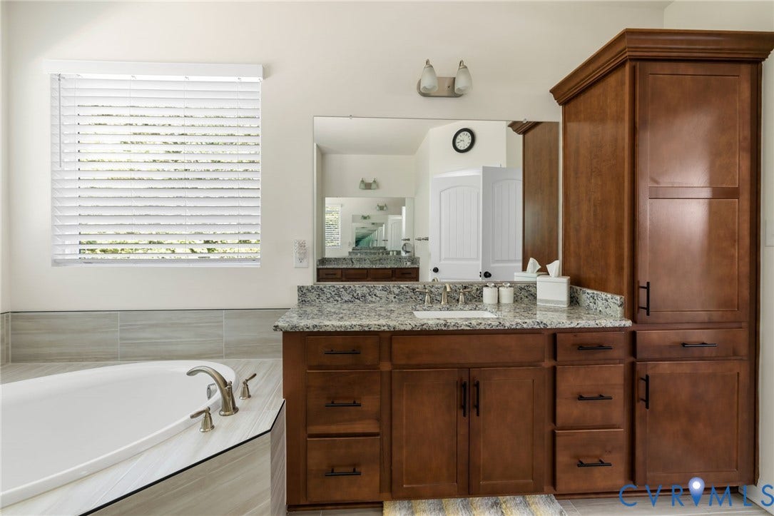 Built-in soaking tub with deck-mounted faucet, wood-finish vanity with an undermount sink, stone countertop, and a tall wood-finish cabinet