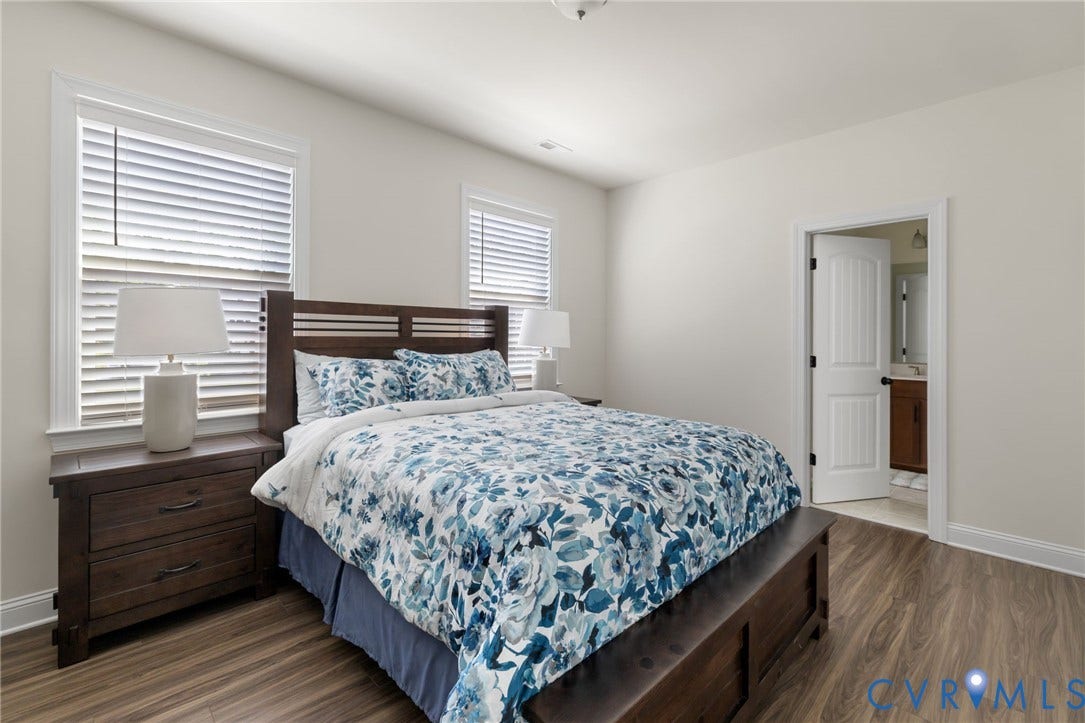 Wood-finish flooring throughout, bright white walls, two windows with blinds, and a paneled interior door leading to an adjoining space