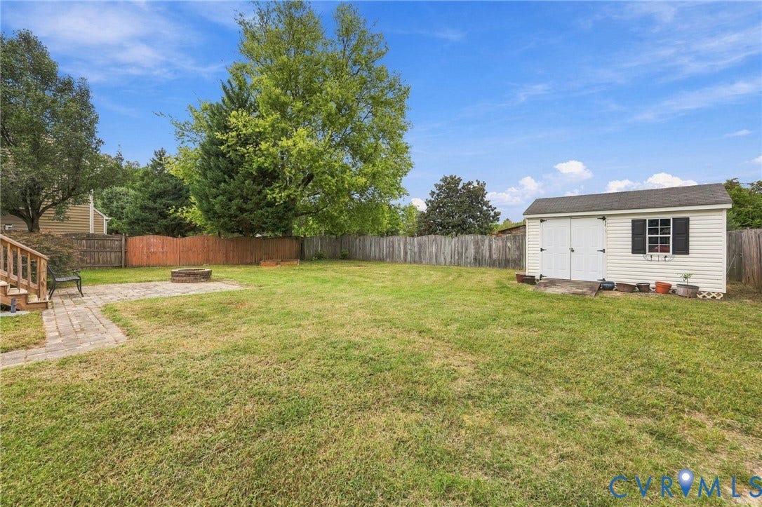 Expansive backyard featuring a paver patio with a fire pit, mature trees, wood fencing, and a white storage shed with a shingled roof