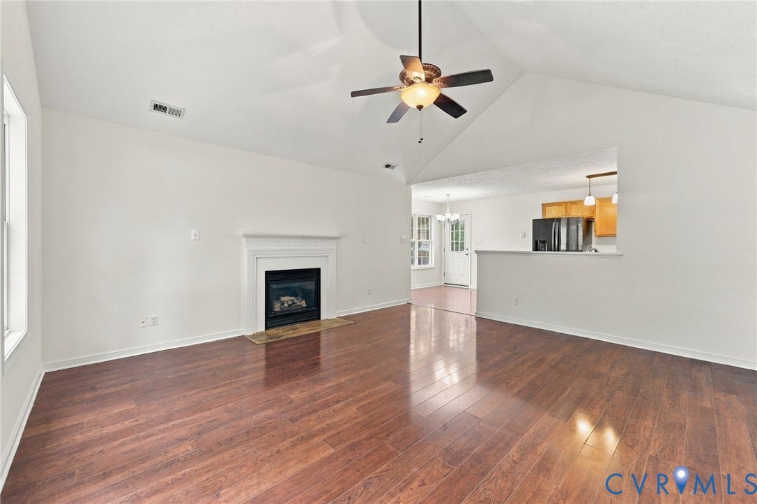 Vaulted ceiling with a ceiling fan, wood-finish flooring, fireplace with a decorative mantel, and a pass-through window to the kitchen